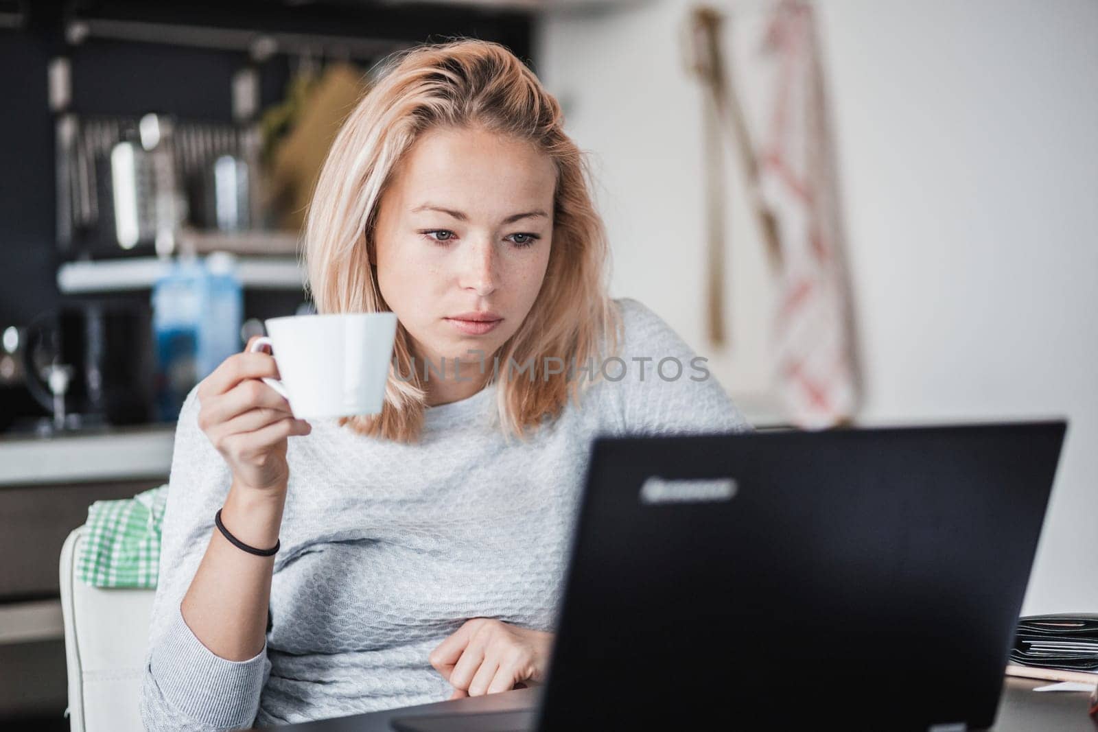 Female freelancer in her casual home clothing working remotly from her dining table in the morning. Home kitchen in the background