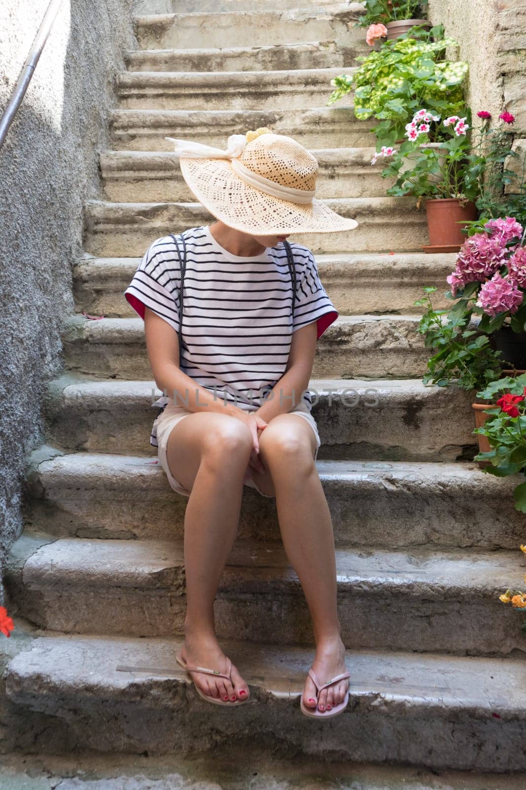 Beautiful female tourist wearing big straw sun hat and shorts sitting and relaxing on old stone house stairs during summer travel Mediterranean costal village on hot summer day.