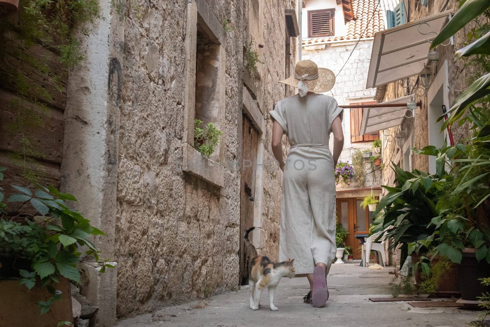 Rear view of beautiful blonde young female traveler wearing straw sun hat sightseeing and enjoying summer vacation in an old traditional costal town at Adriatic cost, Croatia.