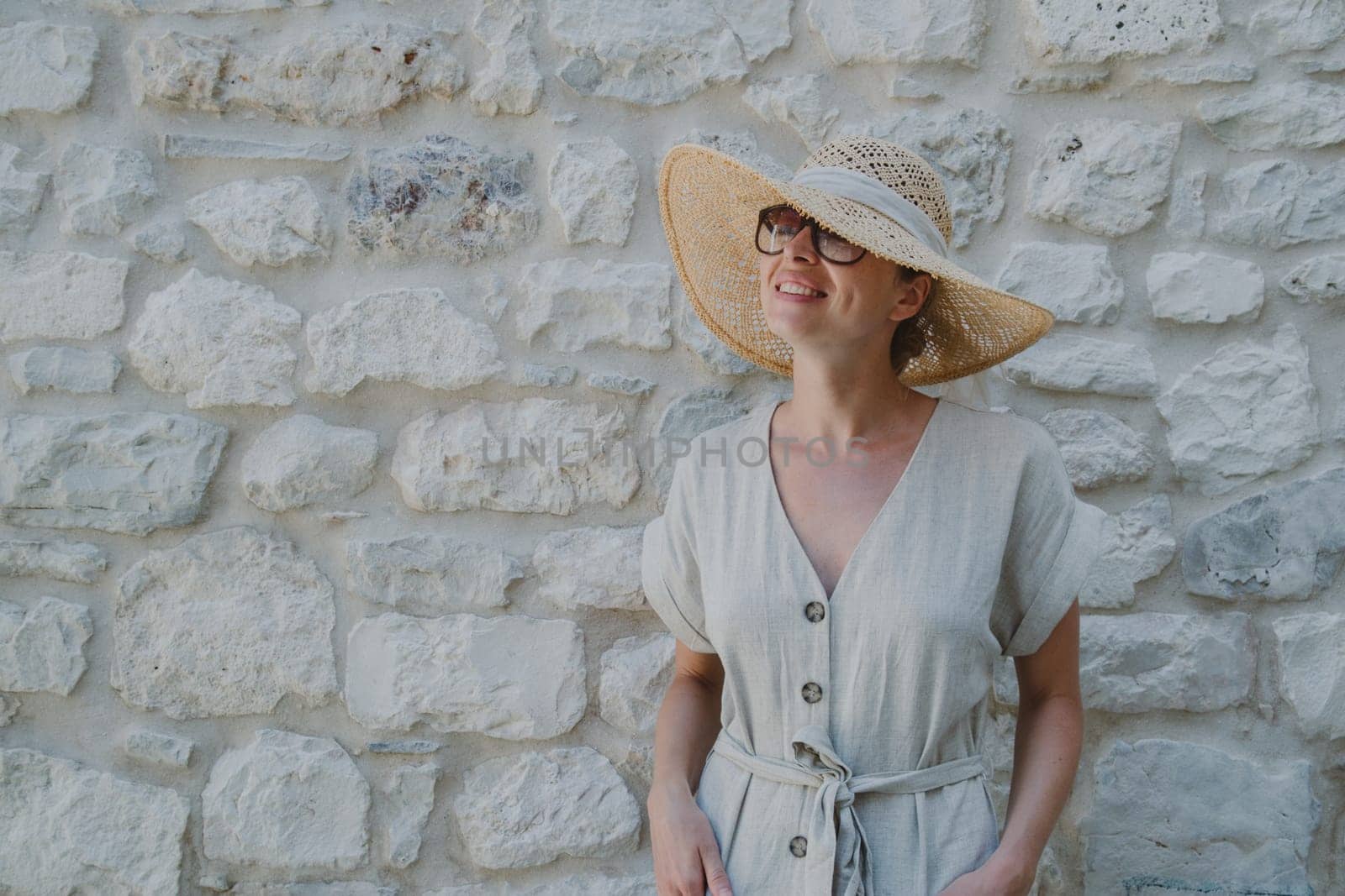 Portrait of beautiful cheerful blonde woman wearing one piece sundress and summer hat, standing in front of old medieval stone wall. Summer vacation portrait concept. Copy space.