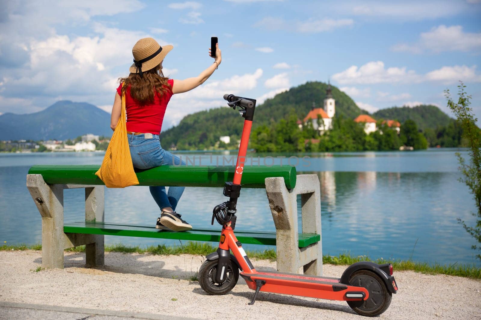 Female traveler taking selfie , sitting at beanch, using electric scooter as modern ecological mean of transport while exploring Bled,Slovenia. Green eco energy concept with zero emission