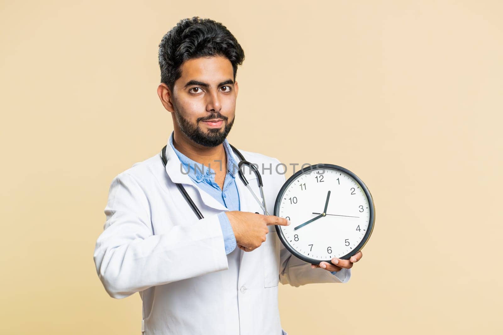 It is your time to heal. Indian young doctor cardiologist man showing time on wall office clock, ok, thumb up, approve, pointing finger at camera. Arabian apothecary pharmacy guy on beige background