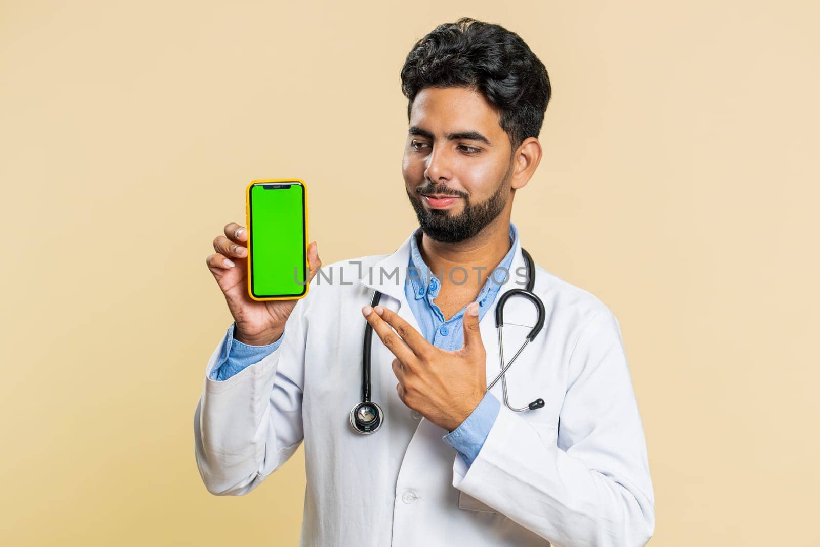 Indian young doctor cardiologist man hold smartphone with green screen chroma key mock up recommend good application promotional sale offer, phone number. Arabian apothecary guy on beige background