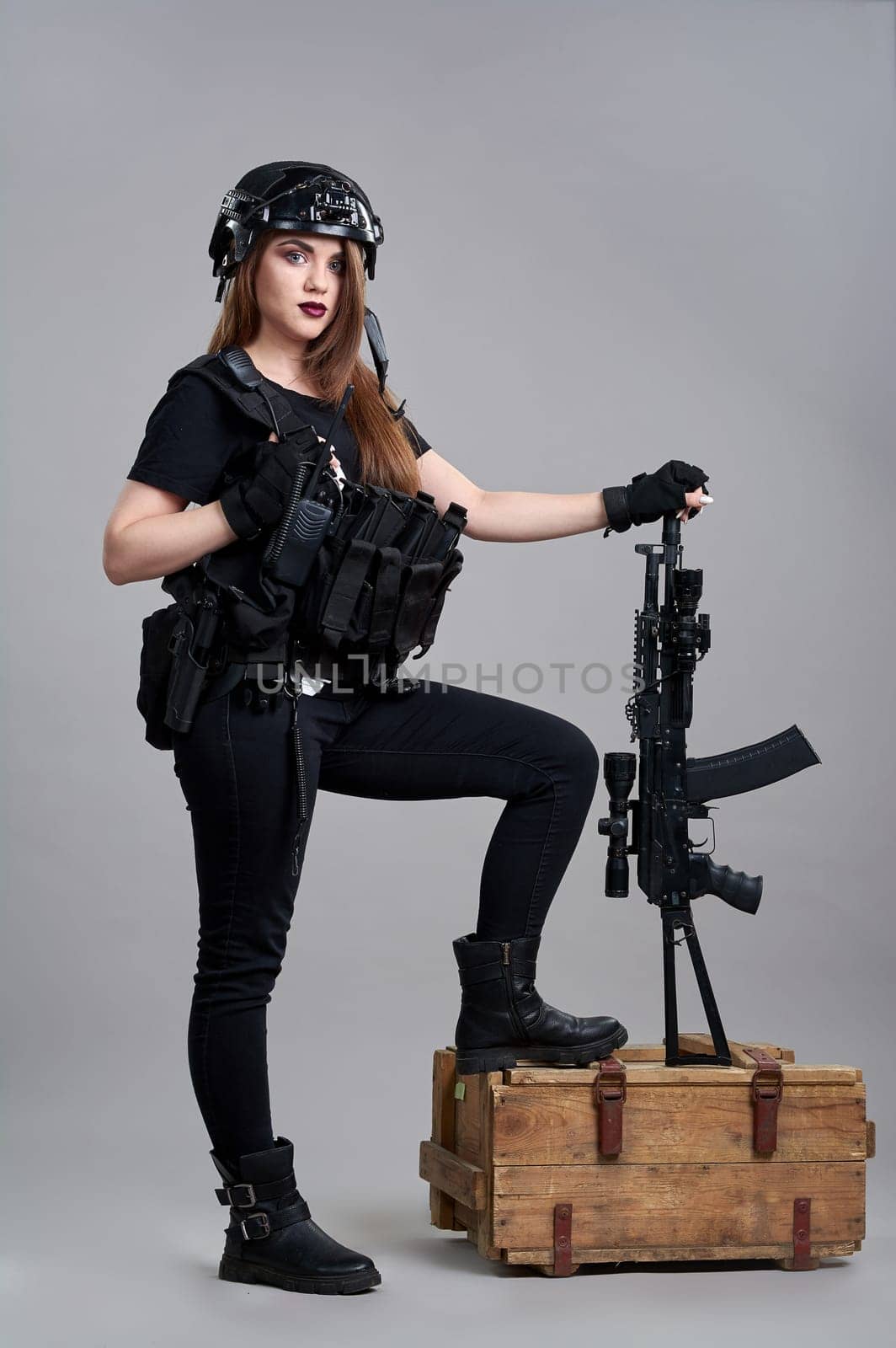 A young woman with a submachine gun leaning on a box of ammunition on a gray background. High quality photo