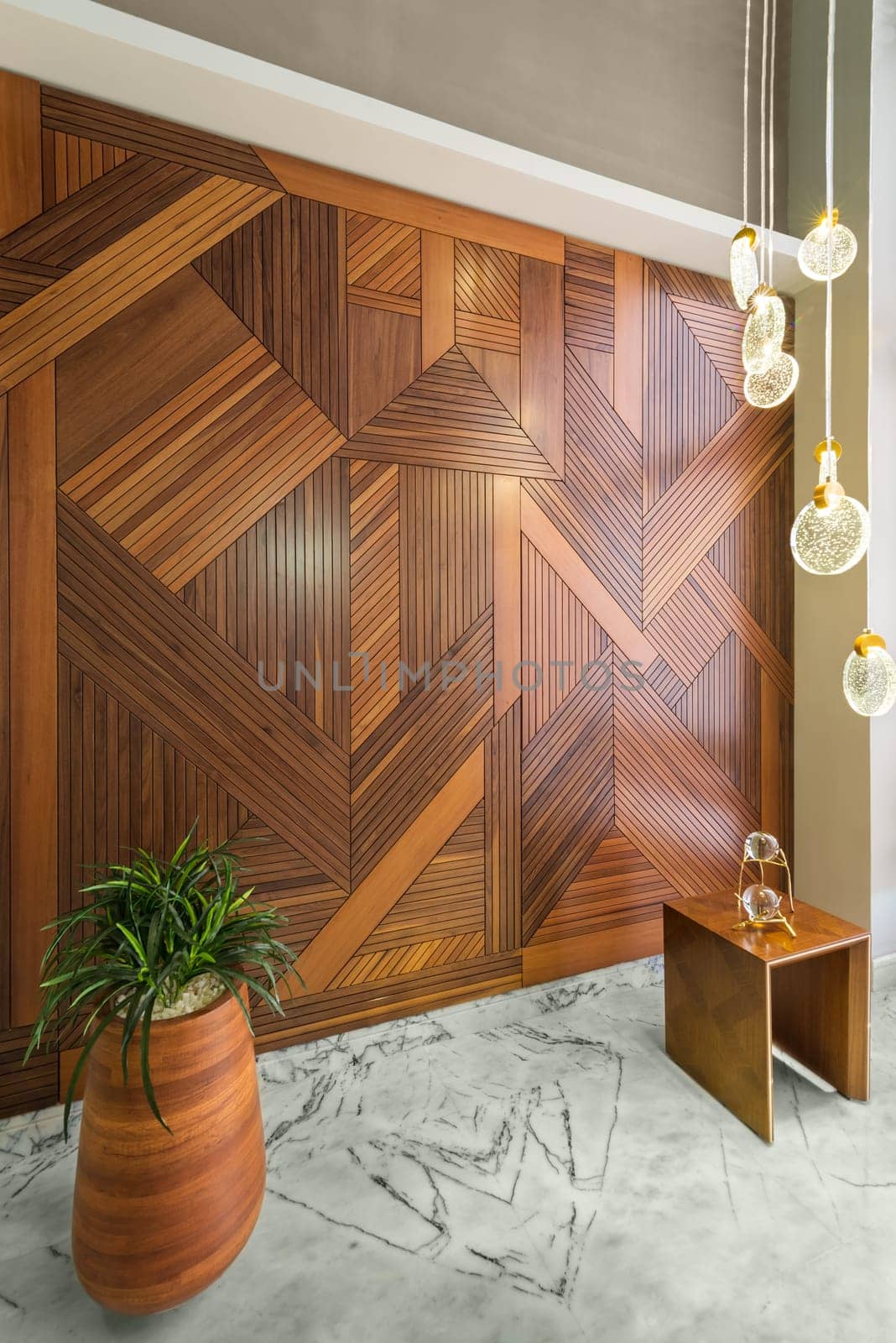 Angled view of hall with wood cladding wall decorated with geometrical patterns, modern glass LED tall chandelier, tall planter with green bushes, and white marble floor in modern house