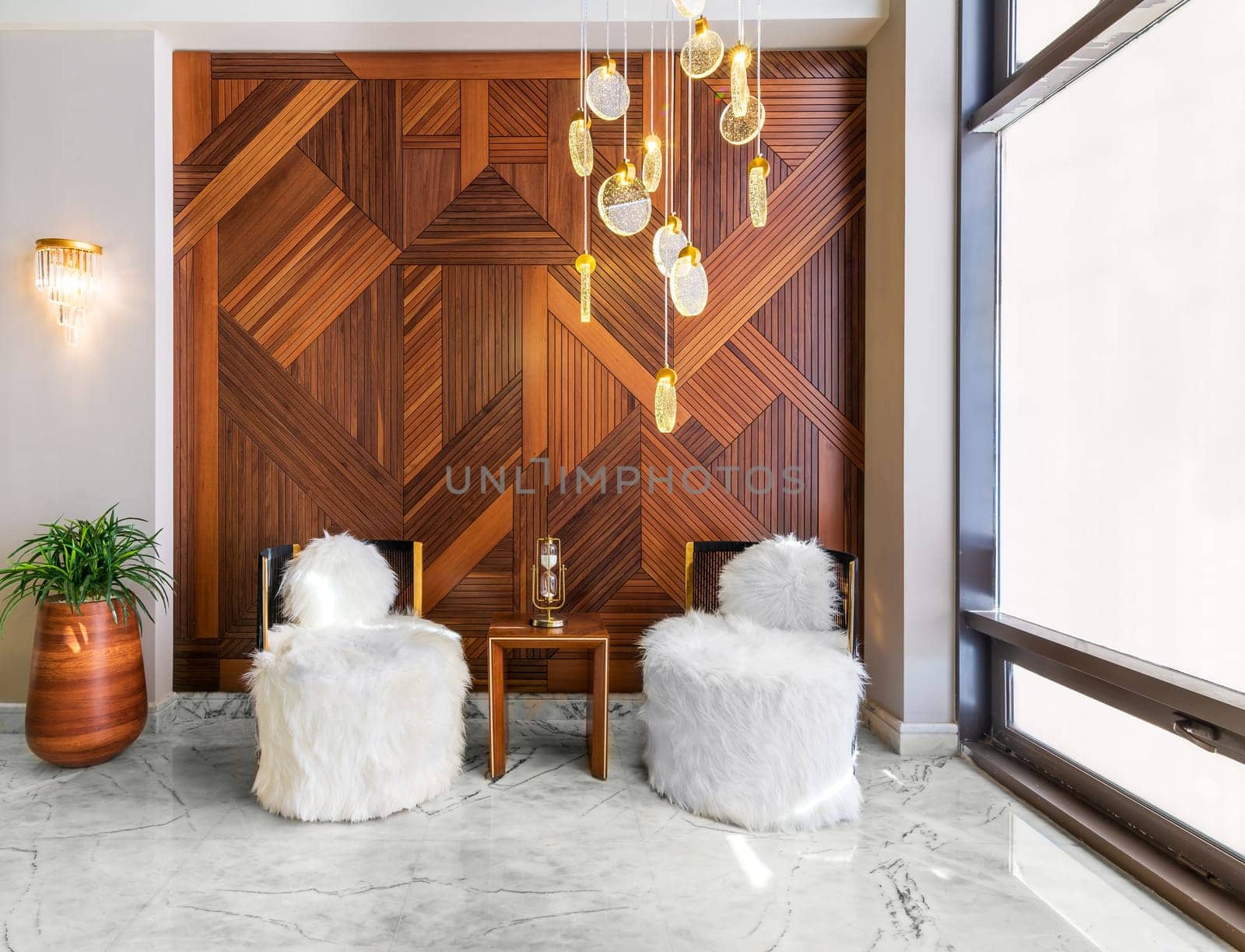 Two modern white feather armless chairs, with small wooden modern table in the middle, and wooden tall planter with green bushes, in a hall with decorated wood cladding wall, and white marble floor