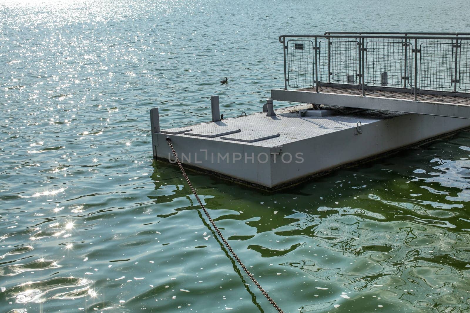 Metal boat pier, on a green water lake, sun sparkling on small waves, chain leading to shore. Brno, Czechia