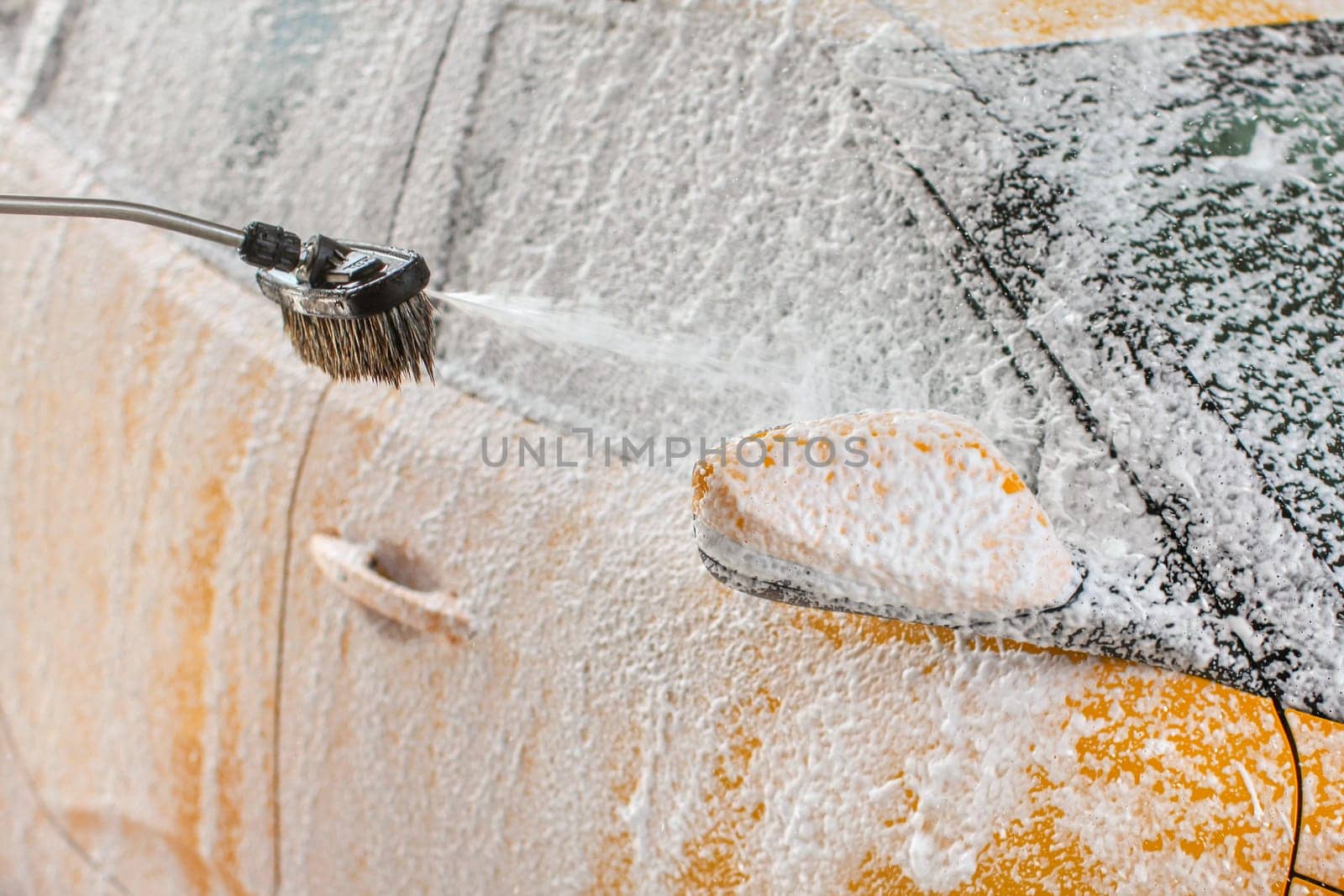 Yellow car being washed in self serve carwash. Shampoo and foam spraying to the side mirror and window.