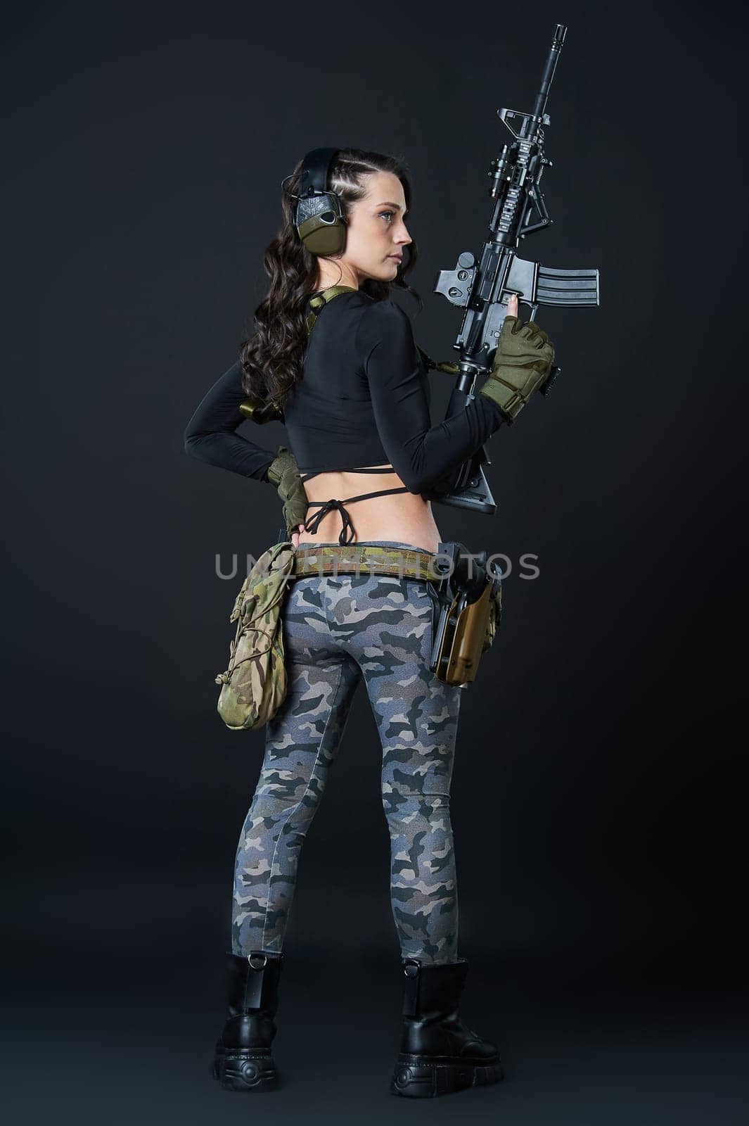 A young woman with a rifle stands with her back against a black background. High quality photo