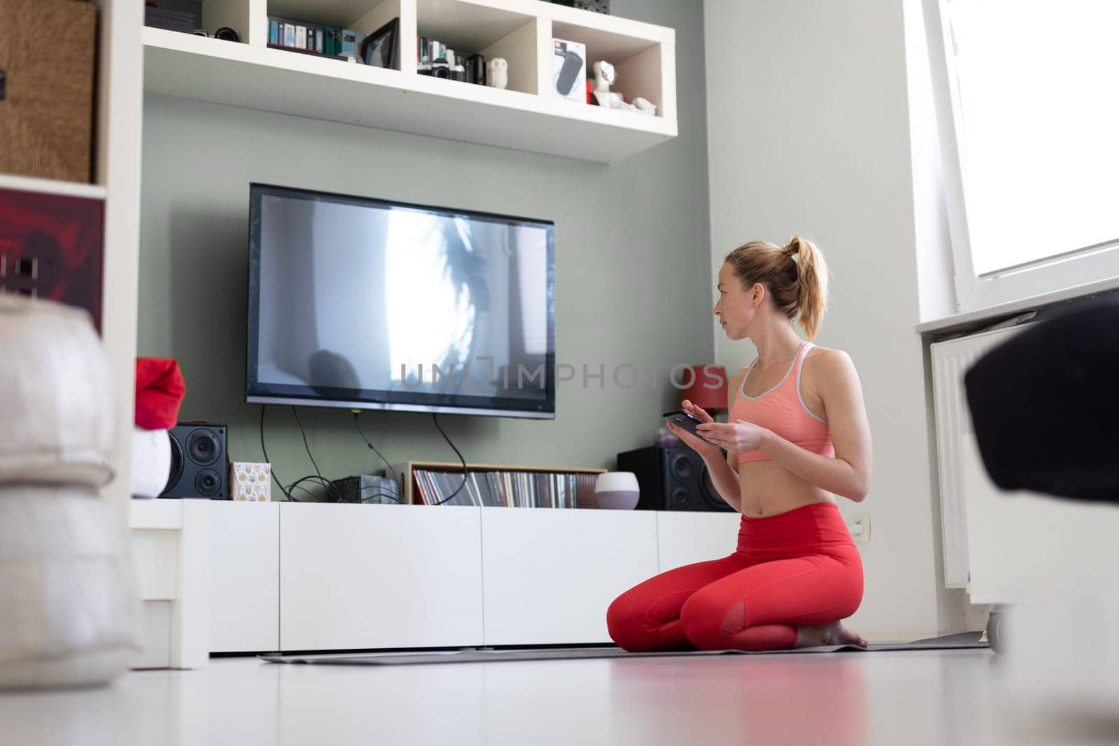 Attractive sporty woman working out at home, doing pilates exercise in front of television in small studio appartment. Social distancing. Stay healthy and stay at home during corona virus pandemic.