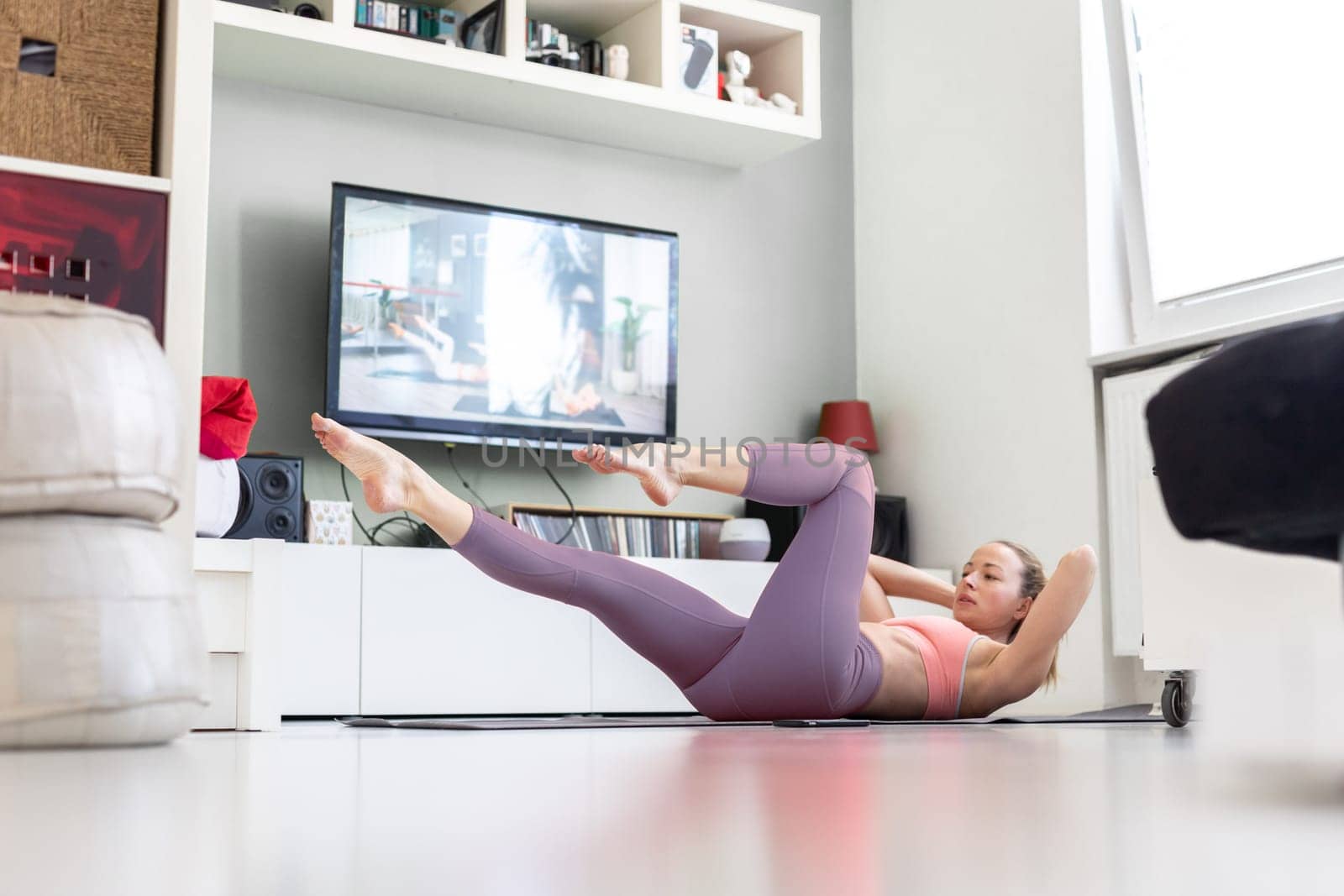 Attractive sporty woman working out at home, doing pilates exercise in front of television in small studio appartment. Social distancing. Stay healthy and stay at home during corona virus pandemic.