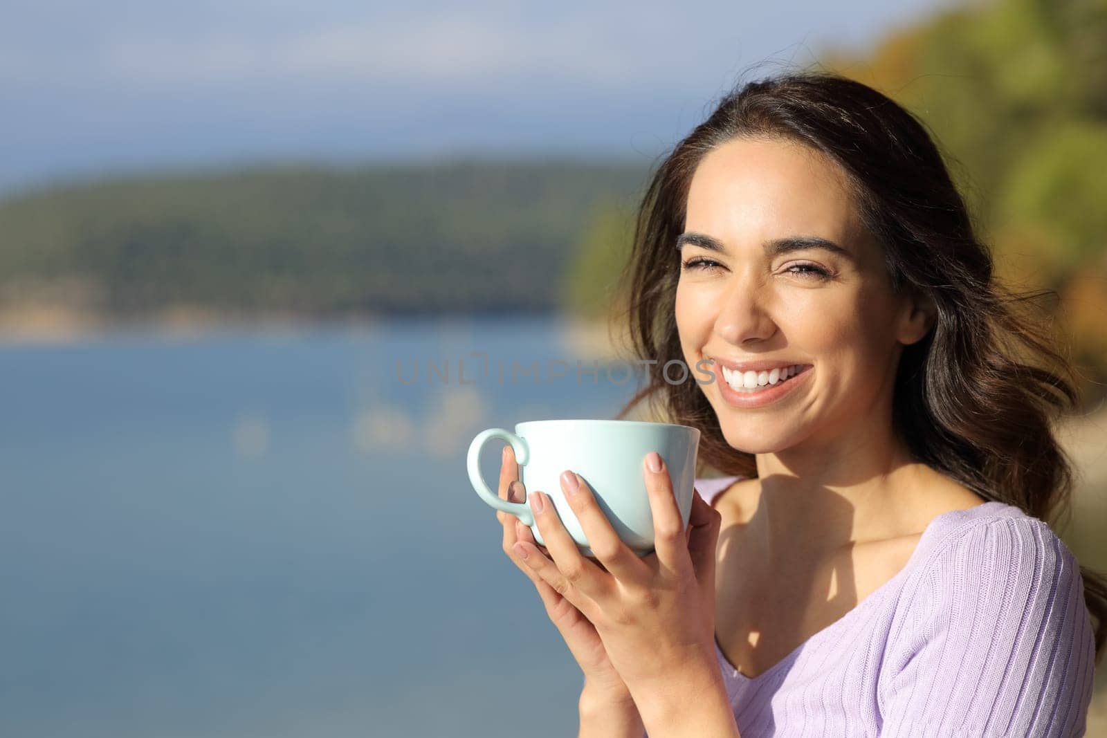 Happy woman holding coffee mug looking at camera in a lake a sunny day