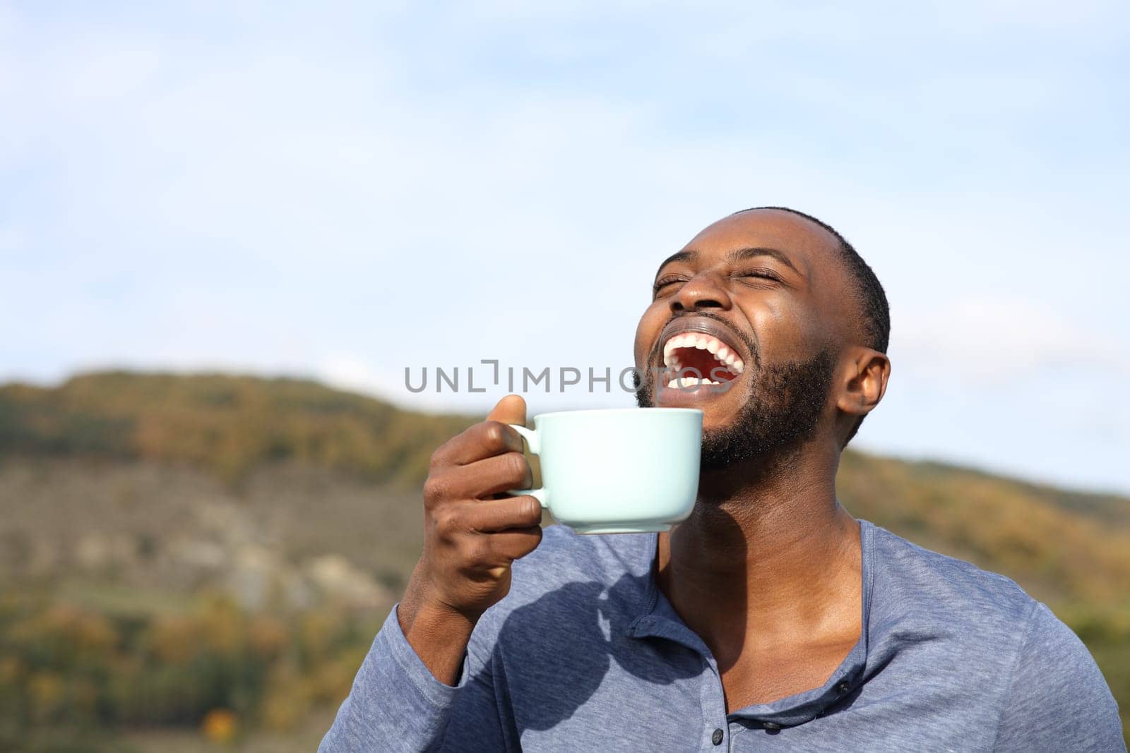 Happy man with black skin laughing and drinking coffee outdoors in the mountain