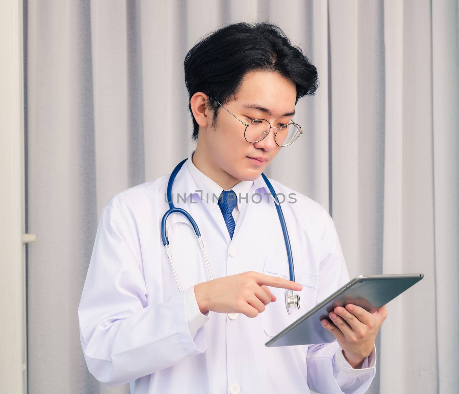 Portrait closeup of Happy Asian young doctor handsome man smiling in uniform and stethoscope neck strap holding and point smart digital tablet and looking to the tablet, healthcare medicine concept