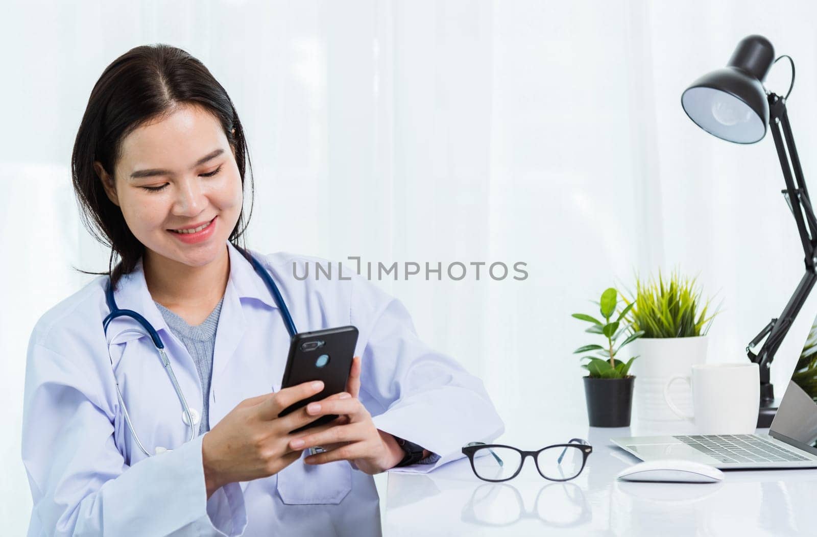 Asian doctor young beautiful woman smiling using working or holding with smart mobile phone and laptop computer at hospital desk office, technology healthcare medical concept