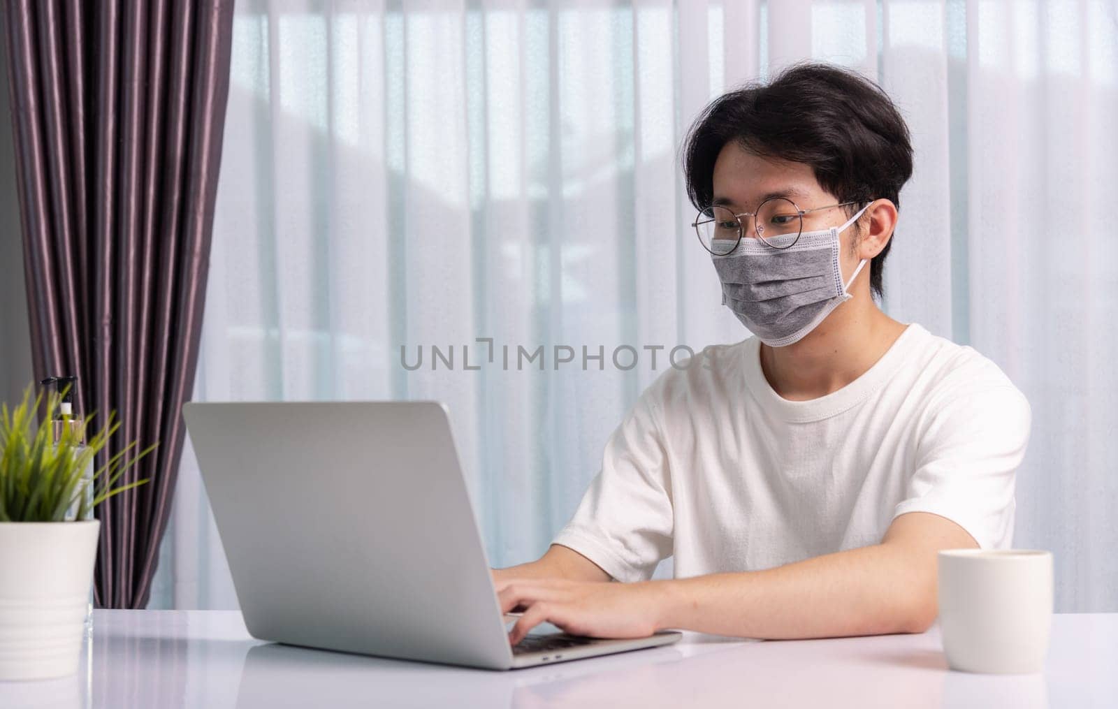Asian business young handsome man glasses, wearing protective mask with disinfectant gel beside he work from home office remote online with laptop computer quarantines disease coronavirus or COVID-19