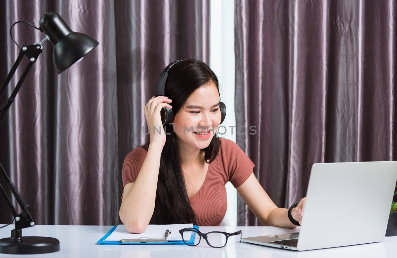 Work from home, Asian young businesswoman smile wearing headphones video conference call or facetime by laptop computer meeting with team on desk at home office
