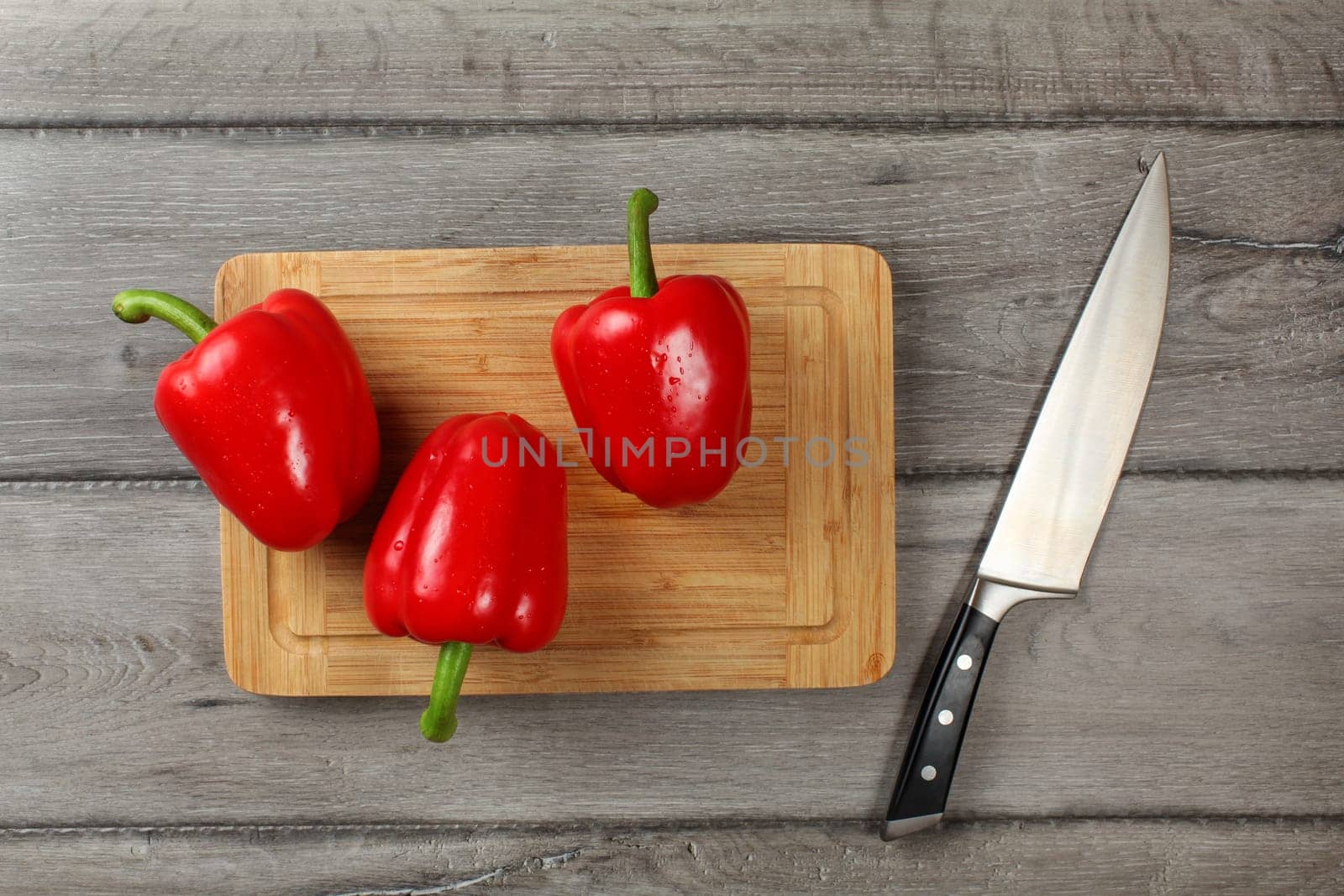 Tabletop view - three whole red bell peppers on chopping board, knife next to it, ready to be cut.
