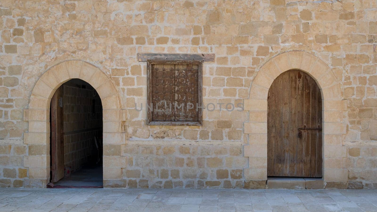 Two weathered wooden arched doors and one closed rusted wrought iron window on bricks stone wall and tiled stone floor
