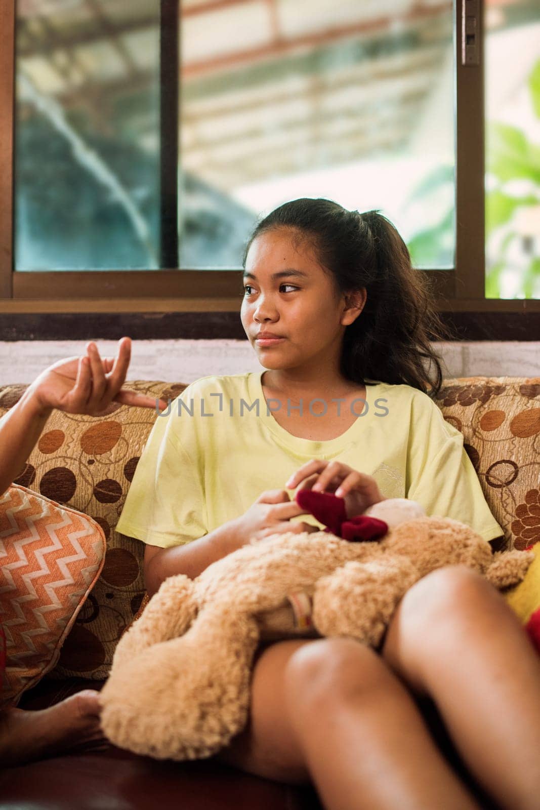 Thai teenager girl sitting on couch with mobile phone and looking to window. Young asian girl looking to side and holding smartphone while sitting on sofa at home. Online education. Using technology. Colourful pillow.