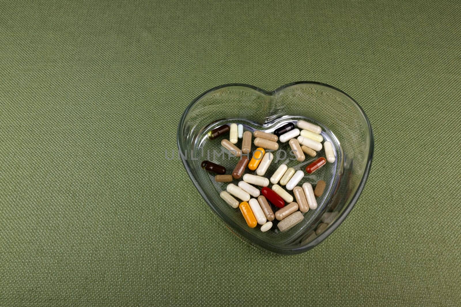A variety of nutritional supplements in a variety of colors and containers like capsules, pills, and softgel in a heart shaped bowl. Health supplements in a heart shaped dish close-up on a tablecloth.