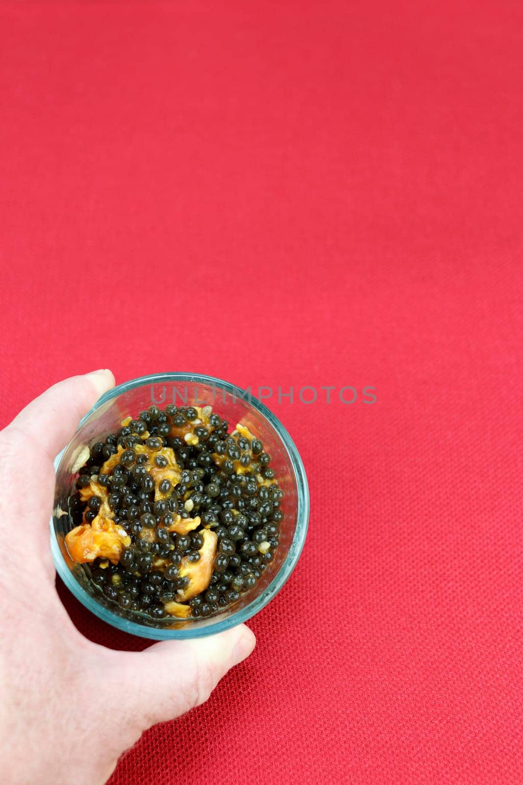 Adult white male holding a small clear glass bowl of papaya seeds and some fruit pulp on a red place mat. White adult male left hand holding a small round bowl of papaya seeds and some papaya fruit.