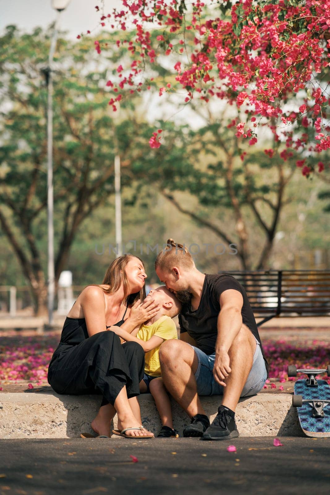 Father and mother is hugging their little son while sitting in skate park with blossom trees around. Concept of family love and care. Outdoor leisure and activity. Happy family.