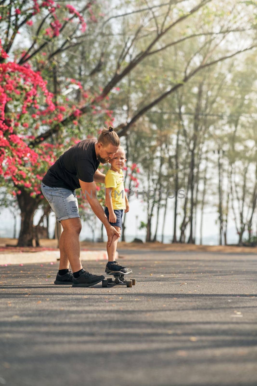 Father holding and training his little son to ride a skateboard in park on spring time. Concept of leisure and outdor family activity. Caucasian man taking care and supporting his kid outdoor.