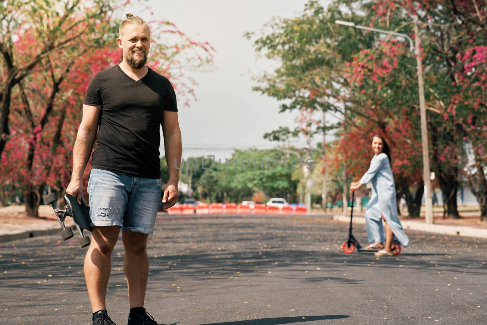 Caucasian happy man with trendy hair style holding skateboard and looking to camera in park while his unfocused wife riding scooter behind. Couple enjoying leisure outdoor.
