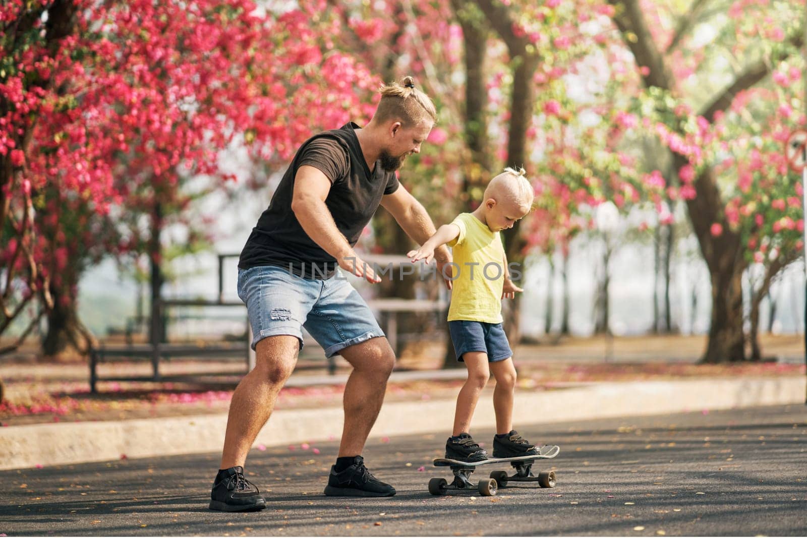 Father holding and training his little son to ride a skateboard in park on spring time. Concept of leisure and outdor family activity. Caucasian man taking care and supporting his kid outdoor.