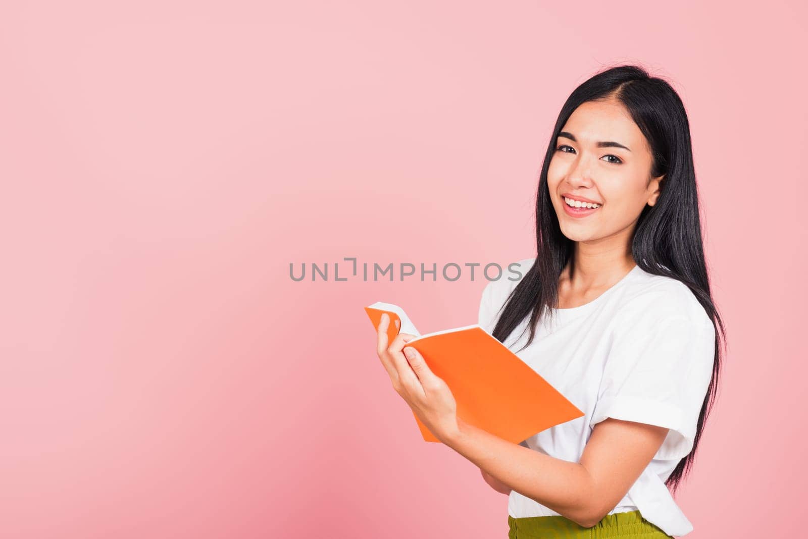 Portrait of happy Asian beautiful young woman confident smiling standing holding orange book open or diary for reading, studio shot isolated on pink background, with copy space, education concept