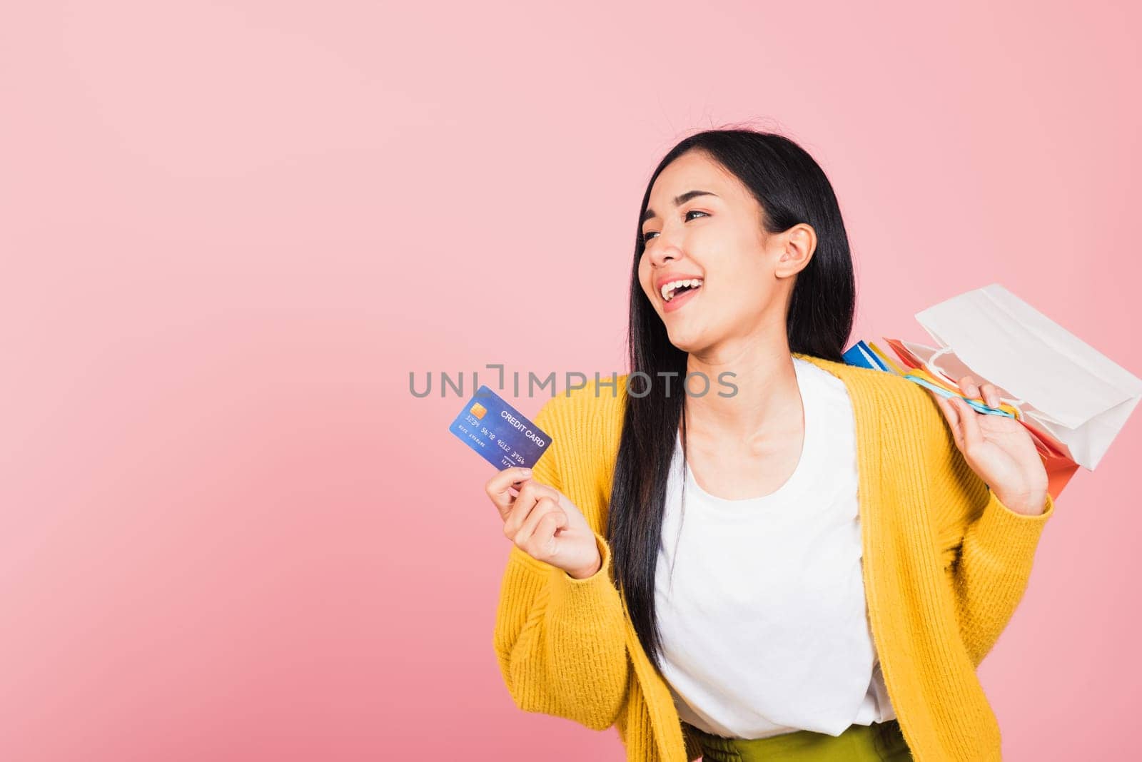 Portrait Asian happy beautiful young woman shopper smiling standing excited holding online shopping bags colorful and credit card for payment on hand in summer, studio shot isolated on pink background