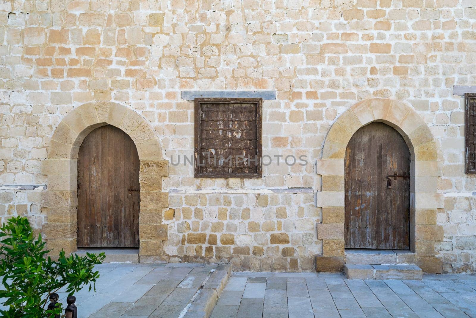 Two weathered wooden arched doors and one closed rusted wrought iron window on bricks stone wall and tiled stone floor
