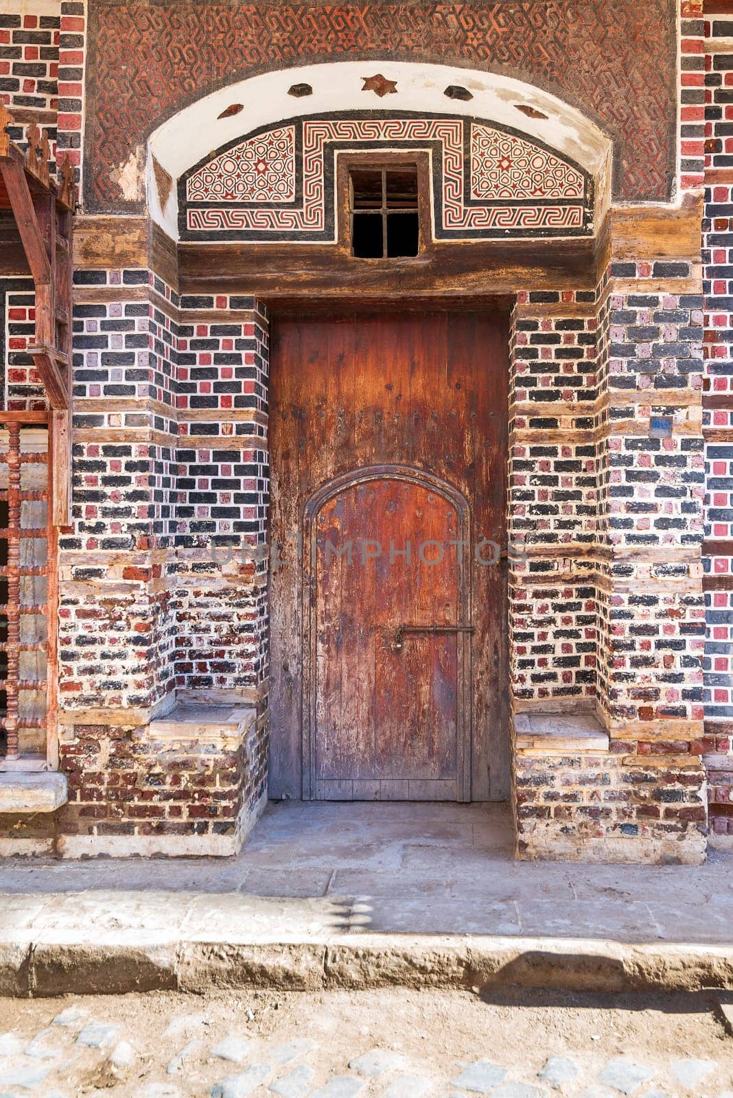 Wooden grunge decorated arched entrance gate with inner small wooden door on wall with black and red bricks with white seam