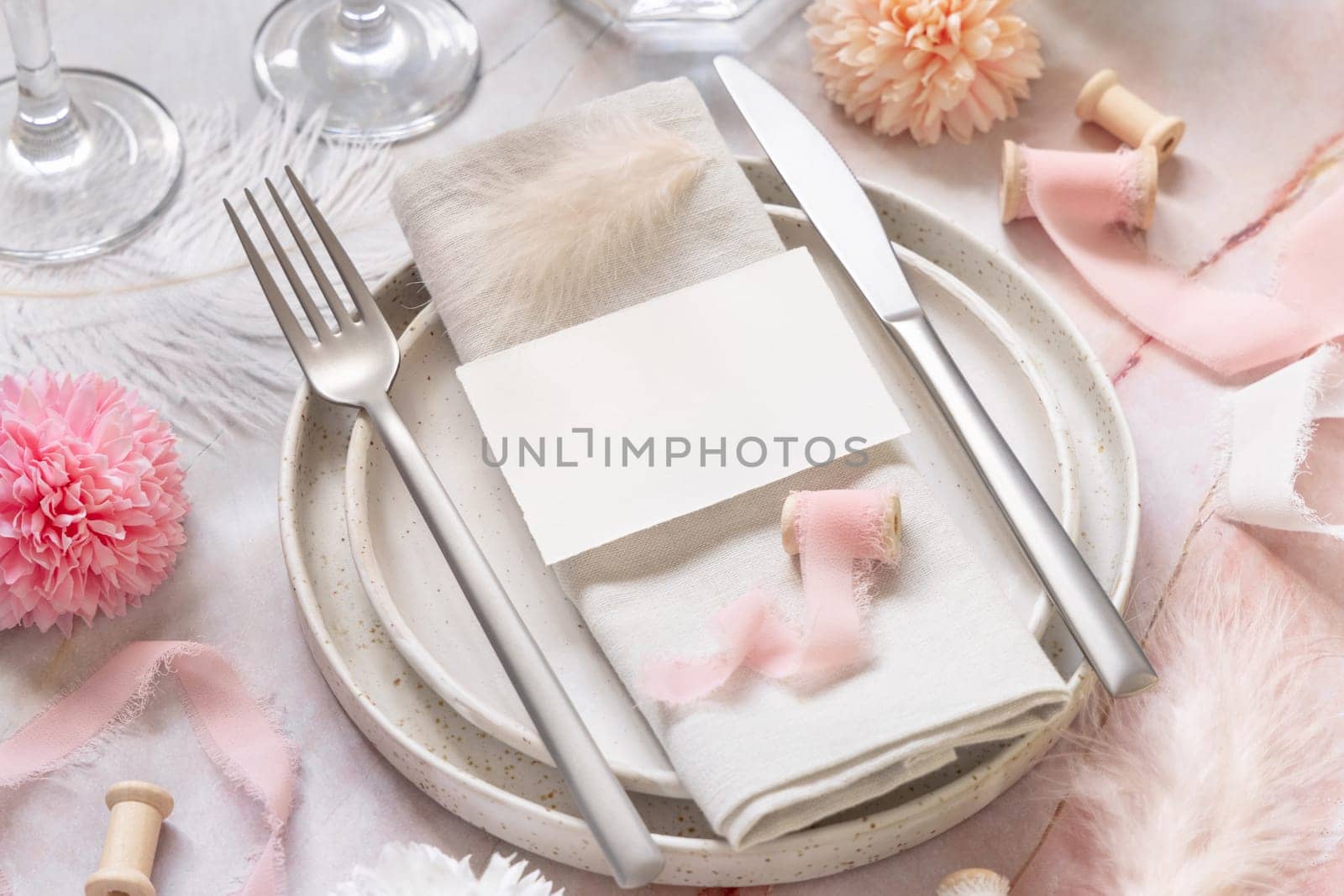 Wedding Table place with a paper card over napkin near flowers, silk ribbons and feathers closeup. Romantic template with blank card on a plate flat lay. Vertical card mockup, copy space