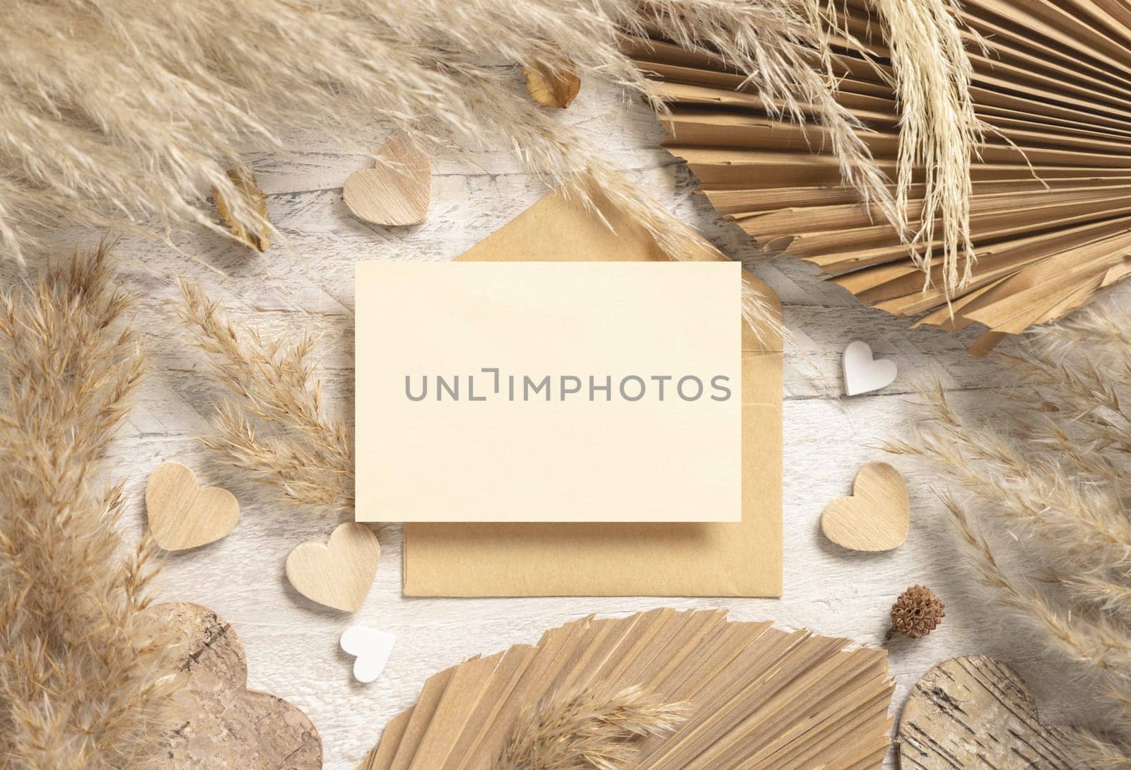 Blank card and envelope on white wooden table near dried pampas grass and hearts top view. Boho scene with horizontal card, flat lay. Bohemian Valentines mockup