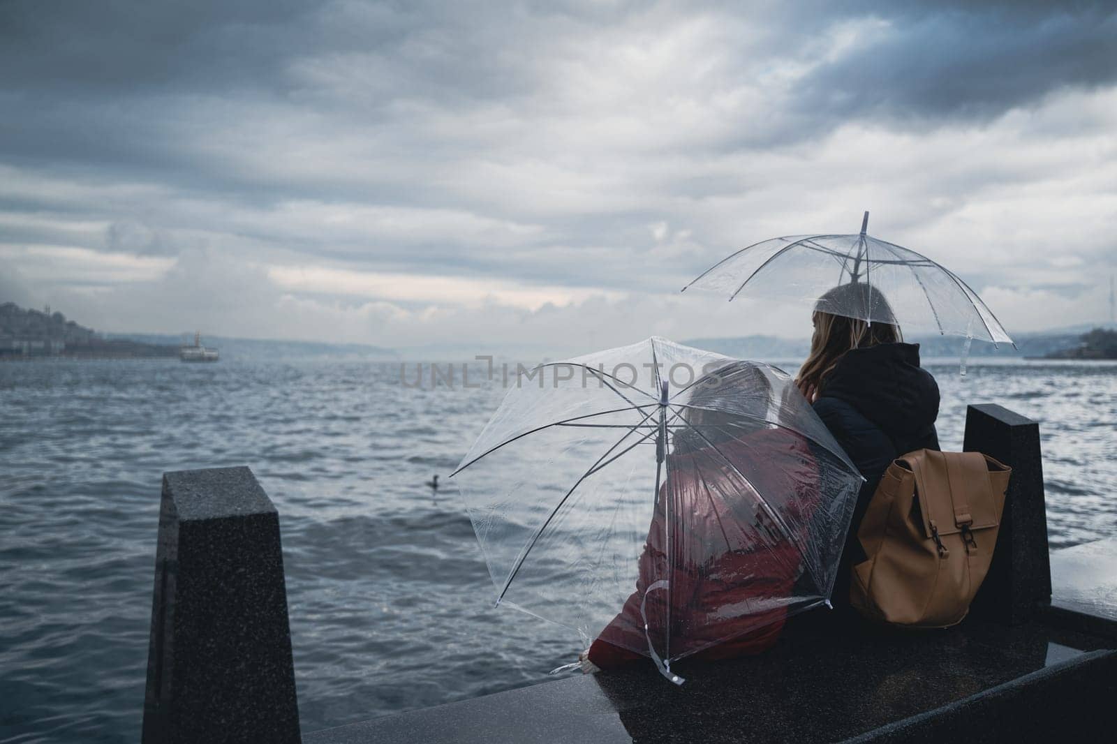 rear view closeup of mother and daughter holding transparent umbrellas sitting on shore and watching the sea on a rainy day with storm clouds above