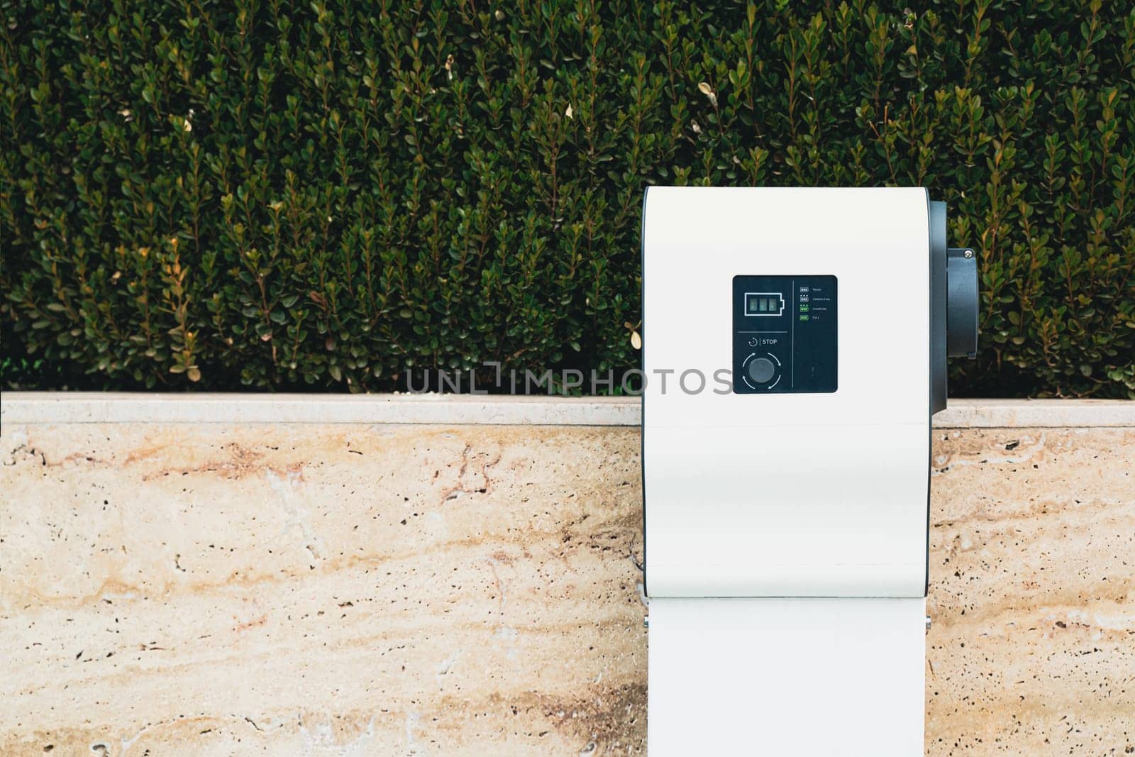 closeup front view of vacant white electric car charger near a stone wall with green vegetation in parking lot