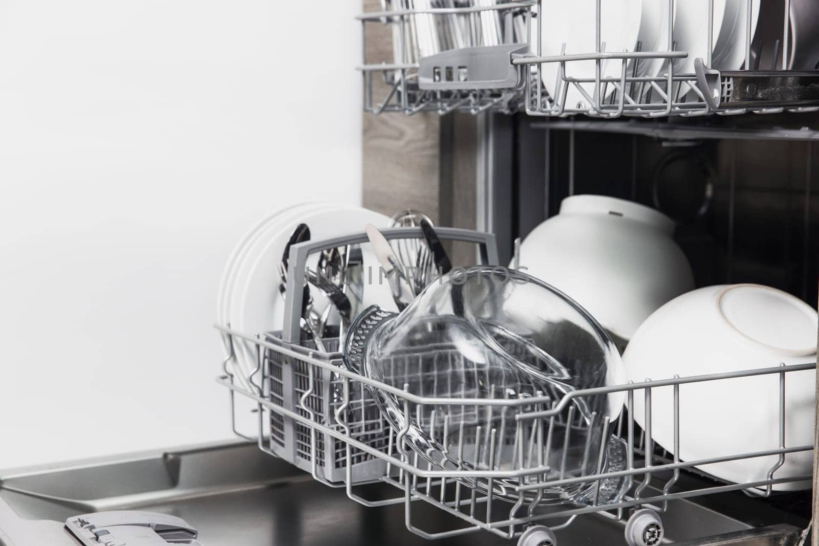 Front view of open automatic stainless steel built-in fully integrated top control dishwasher range machine with clean utensils, cutlery, glasses, dishes, plates inside in modern home kitchen