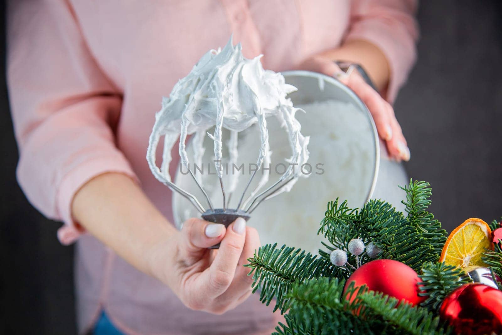 The whisk of an electric mixer in a white mass of meringue with standing peaks is held in the hand of a pastry chef against the background of an iron bowl, the branches of a Christmas tree and New Year's toys are visible from the side. High quality photo
