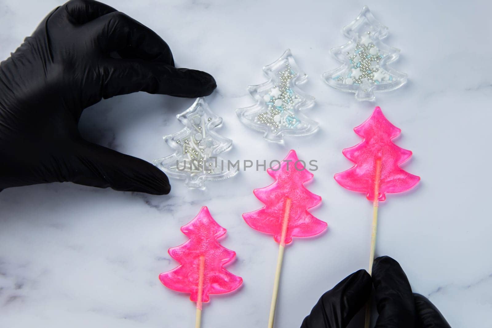Three pink lollipops in the form of Christmas trees lie on white marble, above them are white Christmas trees - lollipops with powder, the extreme ones hold hands in black gloves. High quality photo