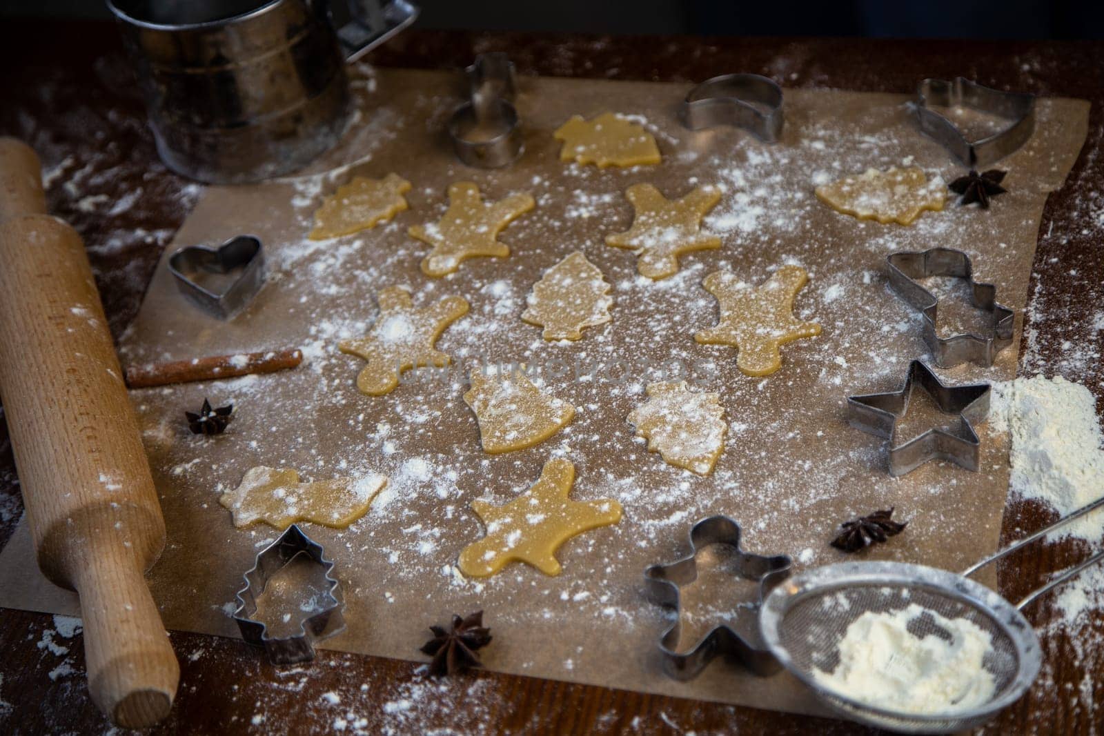 There are ready-made cookies of different shapes on the parchment, a rolling pin, cookie cutters and a sieve with flour are lying nearby, flour is scattered everywhere. High quality photo