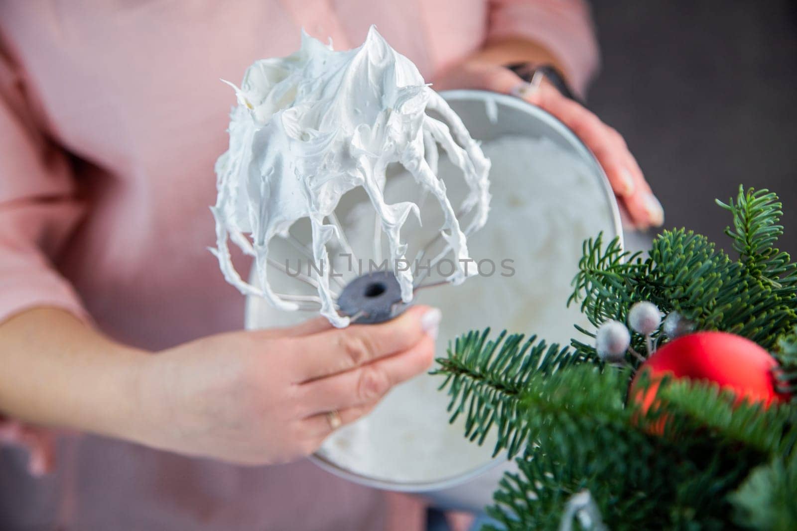 The whisk of an electric mixer in a white mass of meringue with standing peaks is held in the hand of a pastry chef against the background of an iron bowl, the branches of a Christmas tree and New Year's toys are visible from the side. High quality photo