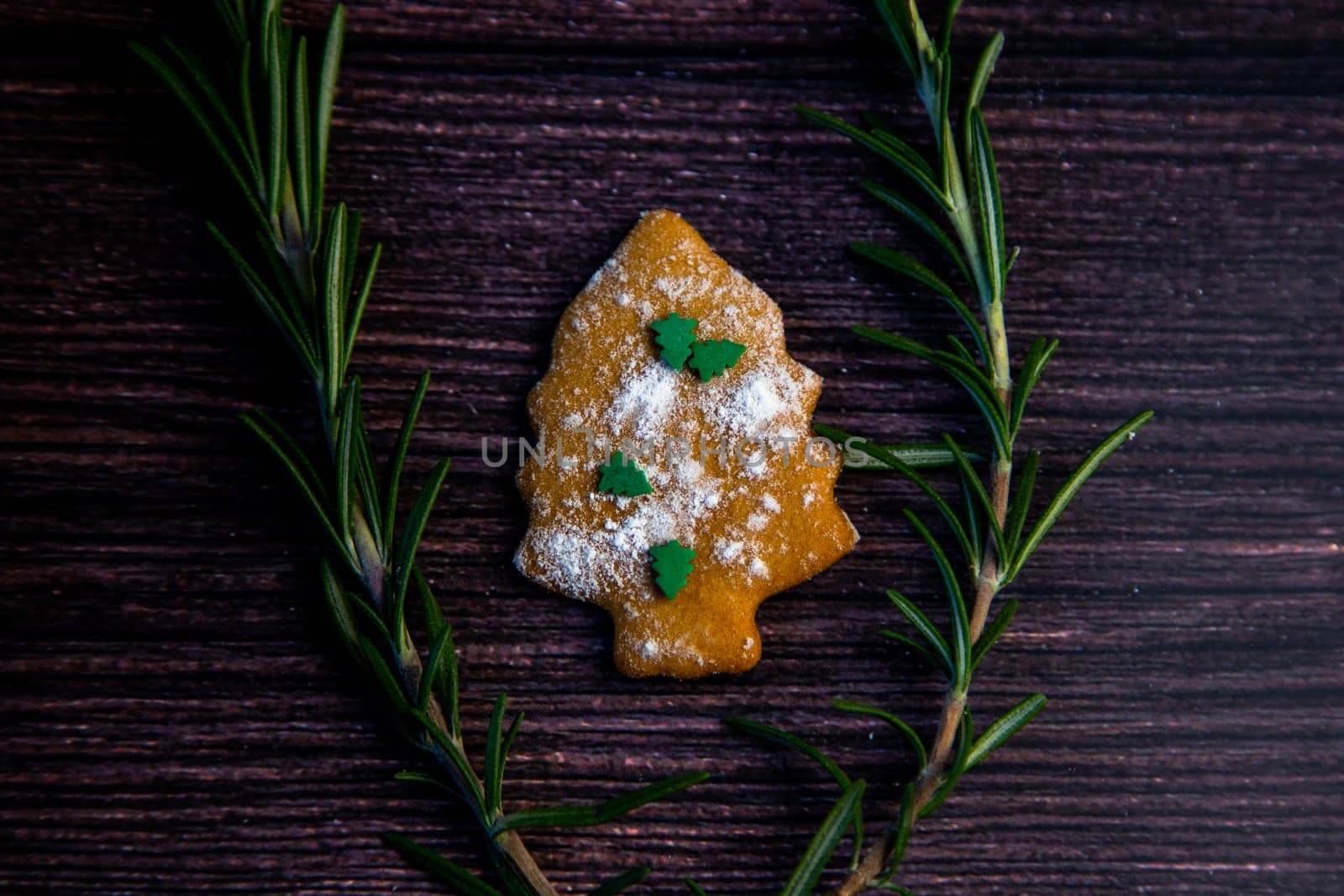 Cookies in the form of a Christmas tree, sprinkled with powdered sugar and small Christmas trees on top, lies on a dark wooden background surrounded by grained rosemary. High quality photo