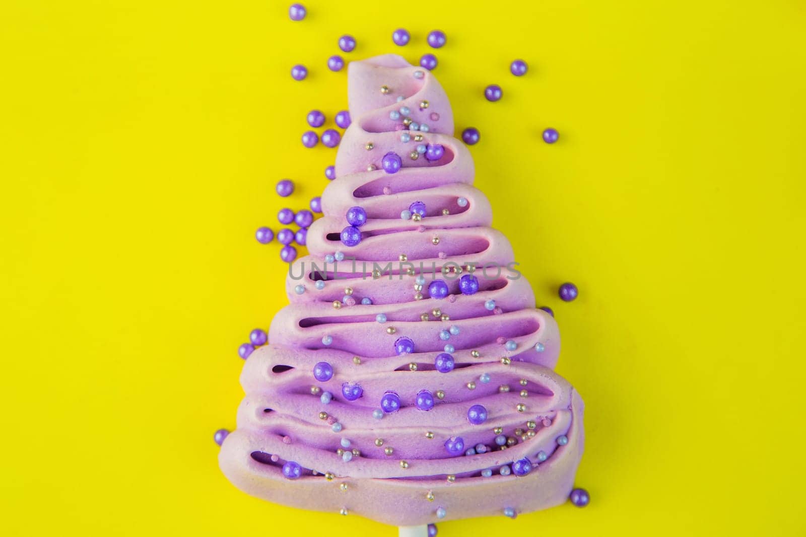 Pale purple Christmas tree made of meringue on a stick, decorated with purple and silver balls on a bright yellow background, purple powder balls are scattered near the tree. High quality photo