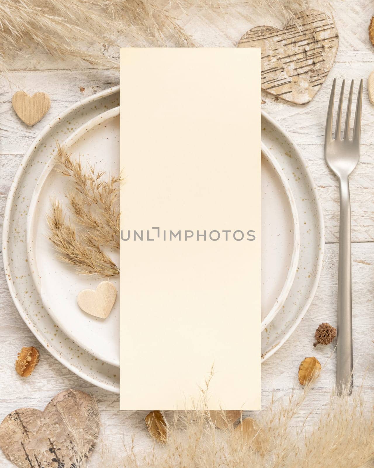 Wedding table place with blank menu card near dried plants, palm leaves, pampas grass and hearts top view. Boho scene with vertical paper card flat lay. Valentines mockup