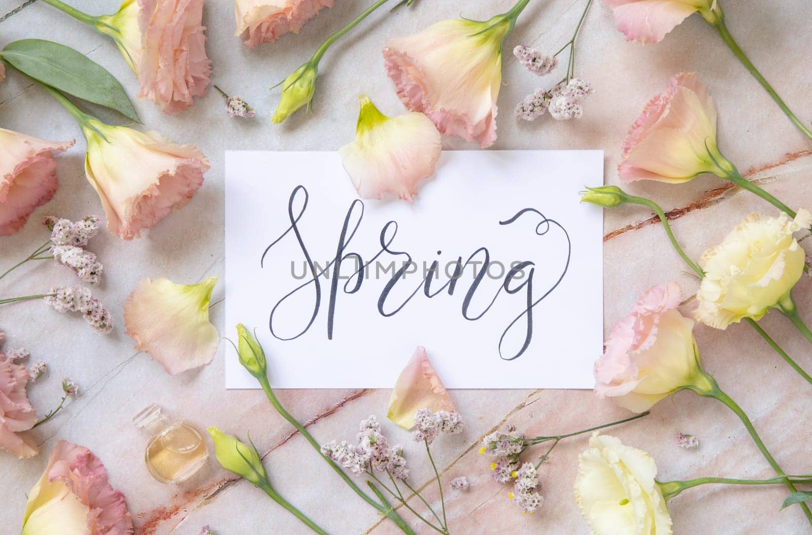 Card with the handwritten text SPRING surrounded by pink flowers, earrings, petals and parfume flacon top view on a marble table. Romantic concept