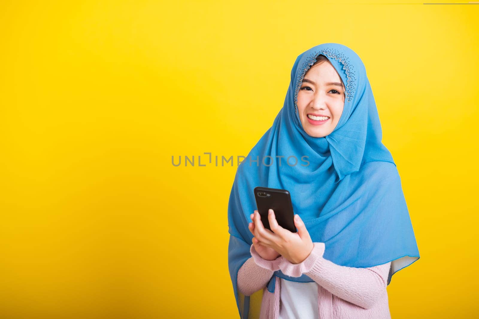 Asian Muslim Arab, Portrait of happy beautiful young woman Islam religious wear veil hijab funny smile she using hold mobile smart phone reading social media isolated on yellow background