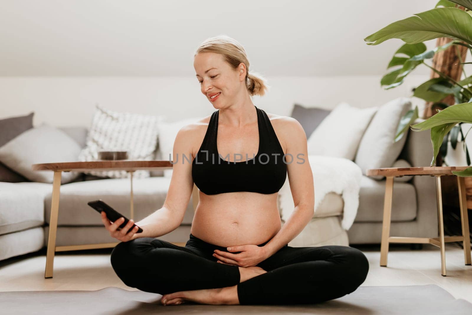 Pregnancy and technoloy supported healthy lifestyle concept. Cheerful happy pregnant woman using smart phone application while exercising on yoga mat on living room floor at home