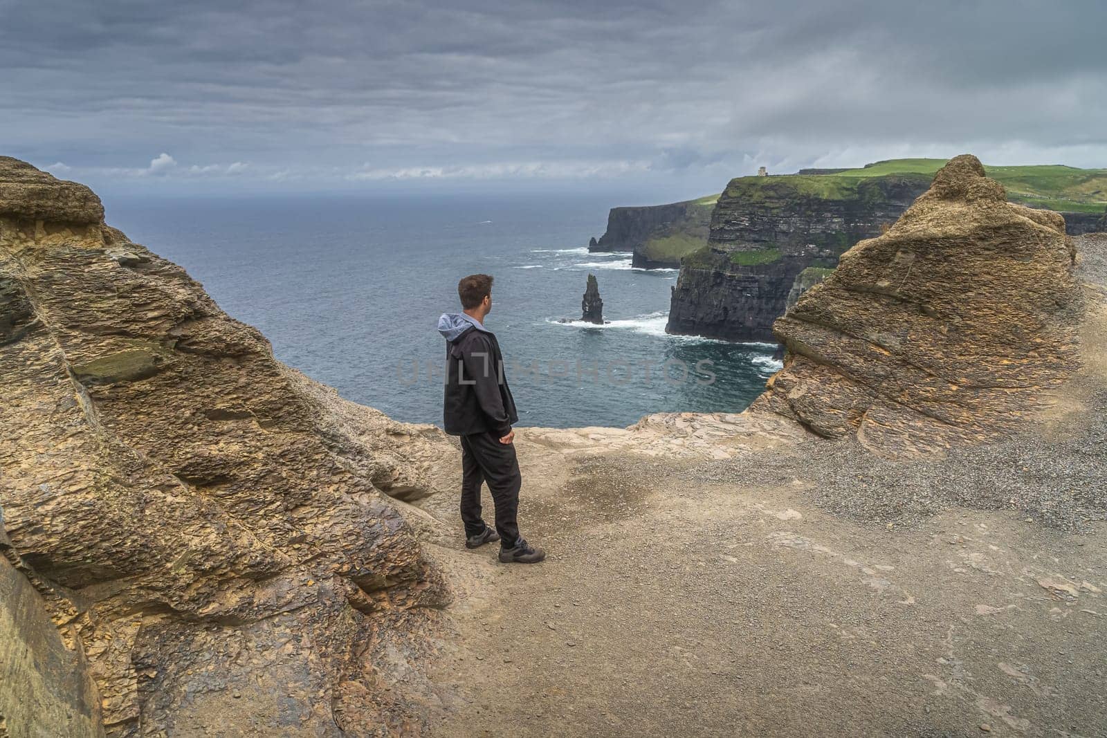Man standing on the edge of iconic Cliffs of Moher and looking at sea stack underneath, popular tourist attraction, UNESCO, Wild Atlantic Way, Ireland